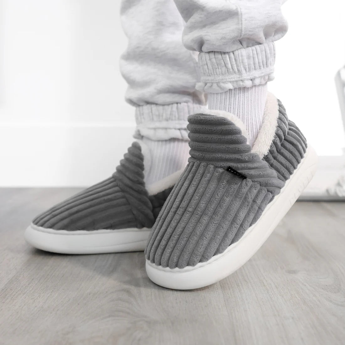 Graue Cord-Hausschuhe an den Füßen einer Person mit weißer Hose